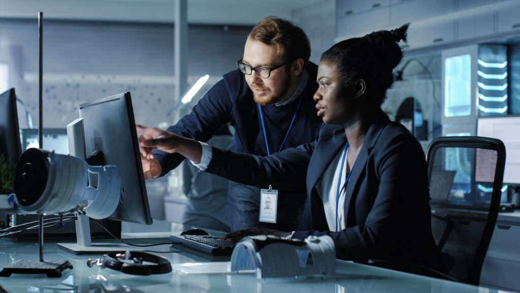 Kubernetes technology concept. Redheaded bespectacled man leans over female colleague as they study desktop monitor.